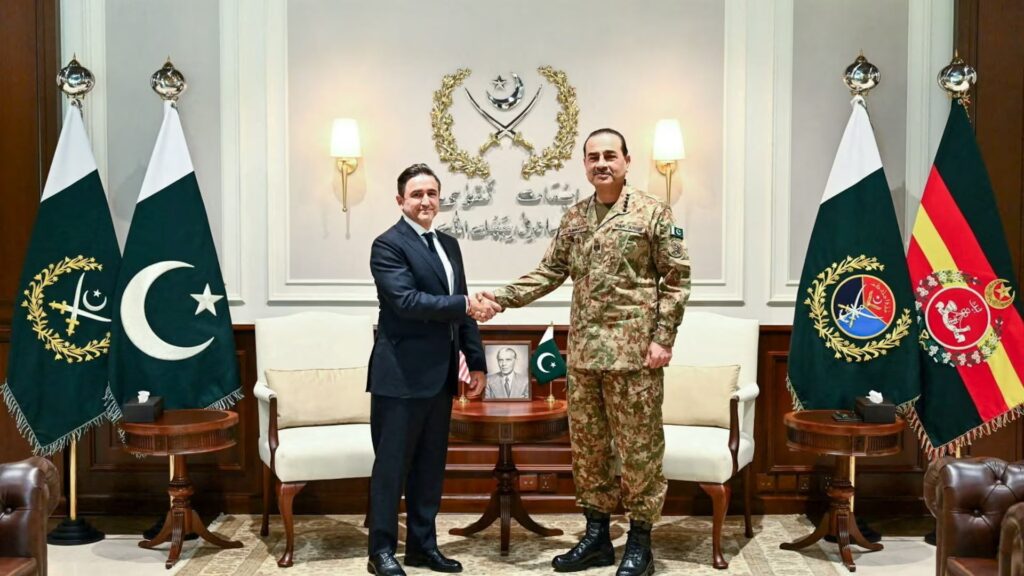 Two men shake hands in a formal office, one in a suit and the other in Pakistani army uniform, flanked by flags on both sides