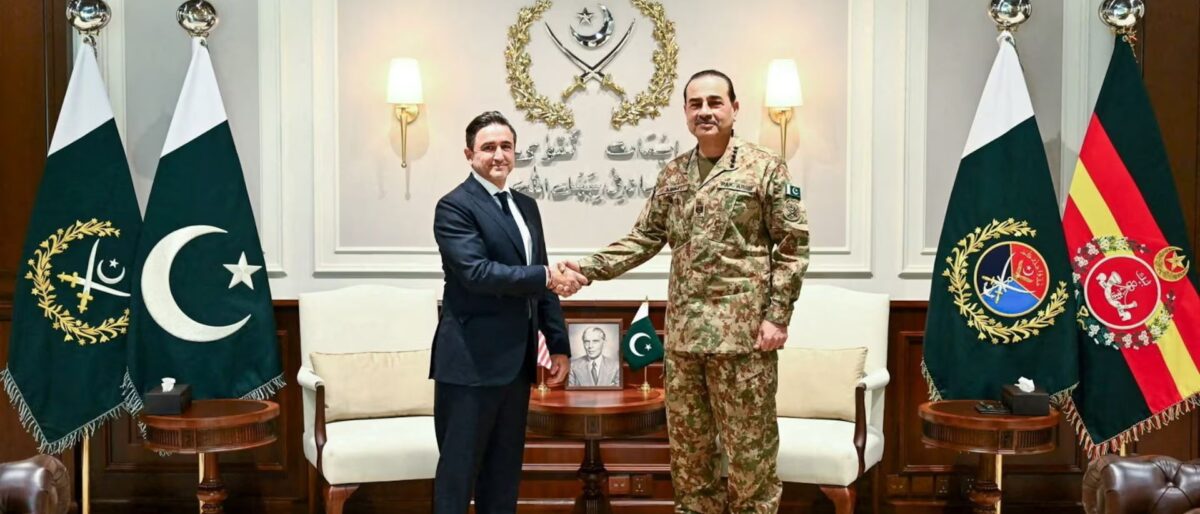 Two men shake hands in a formal office, one in a suit and the other in Pakistani army uniform, flanked by flags on both sides