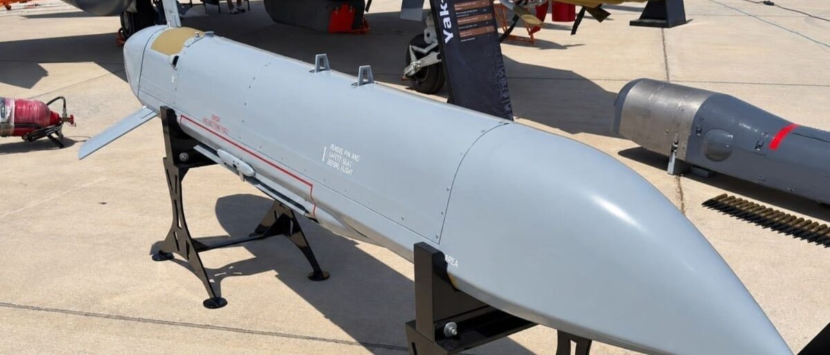 Aircraft exhibit: a long white/gray missiles-like object on a display stand on a tarmac, with parts of an aircraft nearby under a yellow cover in the background.