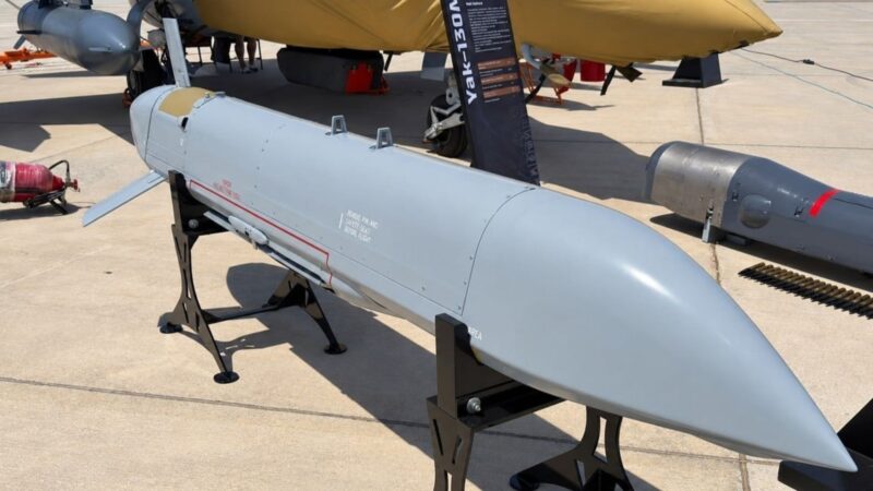 Aircraft exhibit: a long white/gray missiles-like object on a display stand on a tarmac, with parts of an aircraft nearby under a yellow cover in the background.