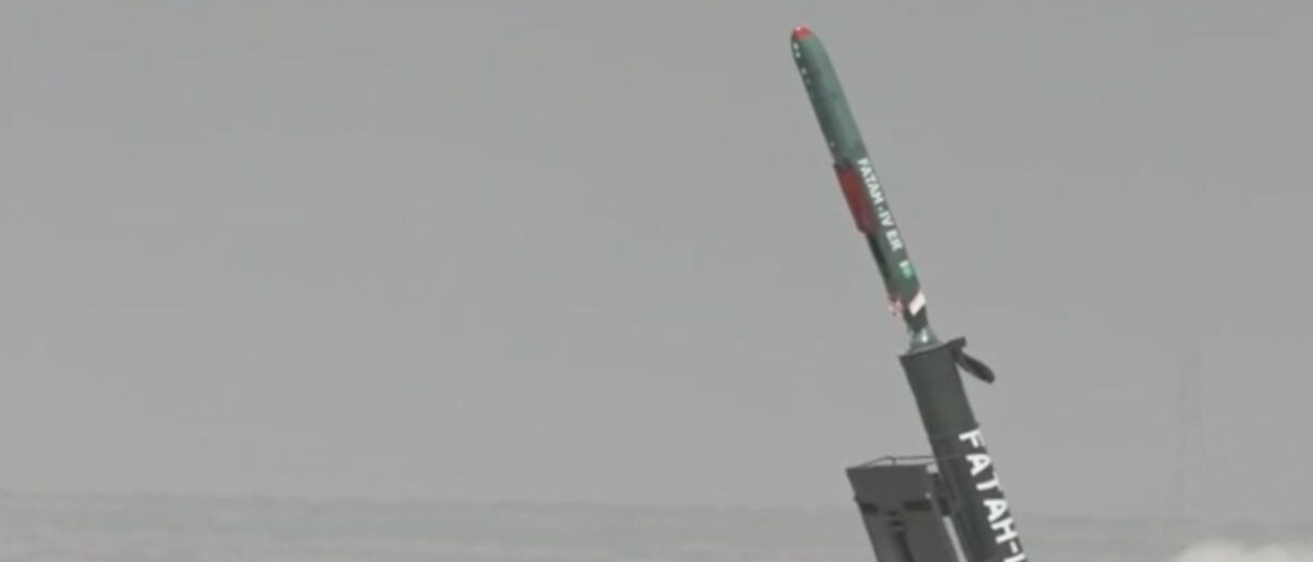 Rocket booster launching from a platform at sea, tilting upward with exhaust plume and dust surrounding it.