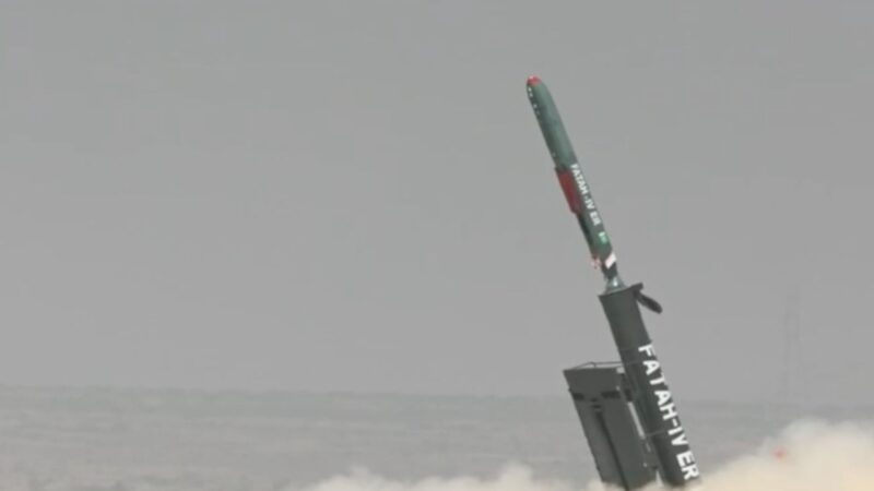 Rocket booster launching from a platform at sea, tilting upward with exhaust plume and dust surrounding it.