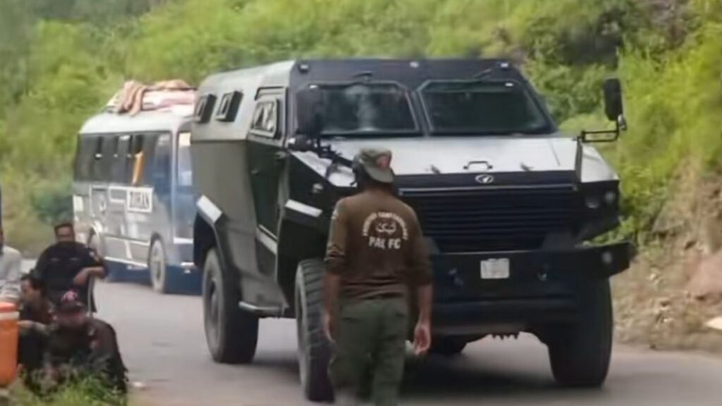 Armored black transport vehicle on a rural road with a man standing nearby and green foliage in the background.