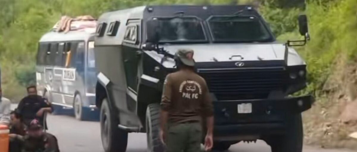 Armored black transport vehicle on a rural road with a man standing nearby and green foliage in the background.