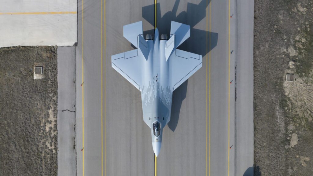 Turkish Aerospace KAAN - Quwa Top-down aerial view of a light-gray fighter jet parked on a paved runway with yellow lines and surrounding terrain on either side.