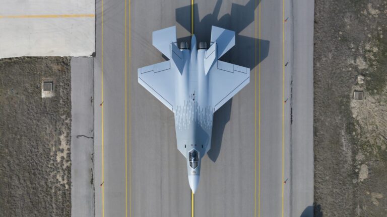 Turkish Aerospace KAAN - Quwa Top-down aerial view of a light-gray fighter jet parked on a paved runway with yellow lines and surrounding terrain on either side.