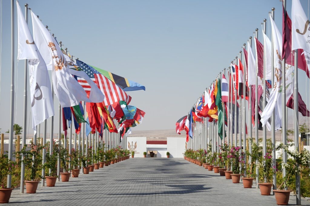 Row of international flags on flagpoles lining a paved walkway toward a white building in a desert setting under a clear blue sky.