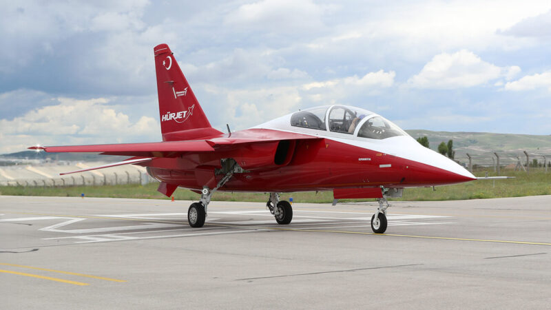 Red and white HÜRJET jet trainer with its landing gear down on an apron